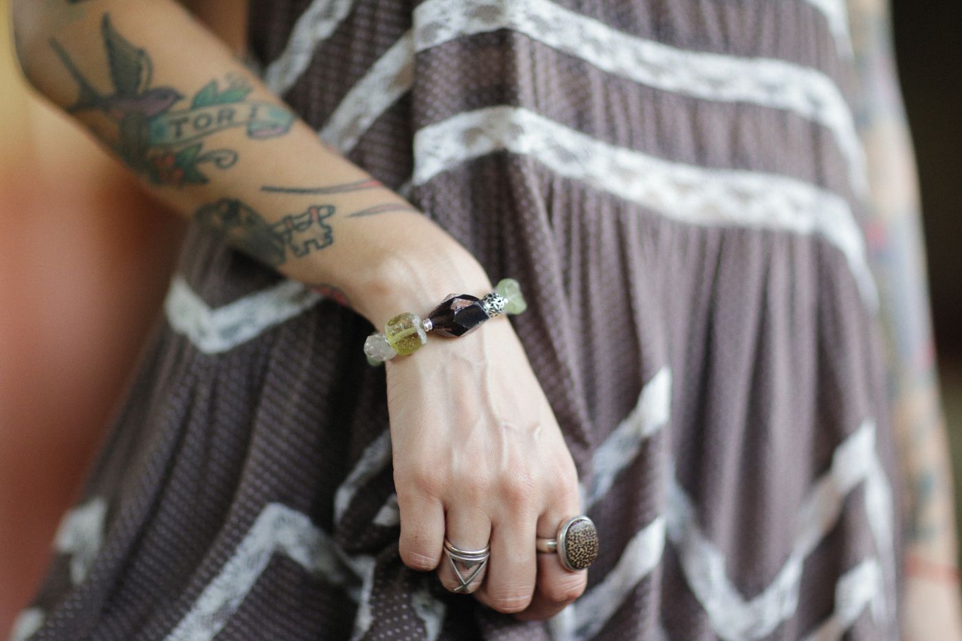 A tattooed arm wearing a gemstone bracelet and rings over a patterned dress